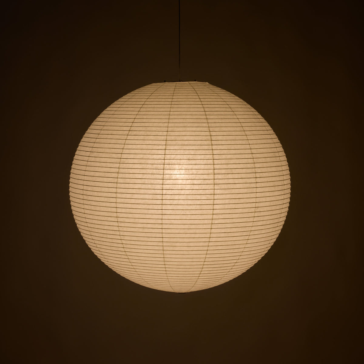A spherical paper lantern pendant light glowing with warm cream light against a dark brown background.