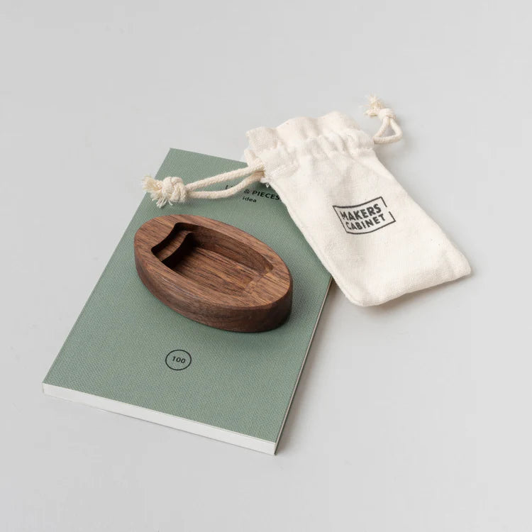 Wooden dish on a green book with a white drawstring bag labeled 'Makers' Cabinet' on a light gray background