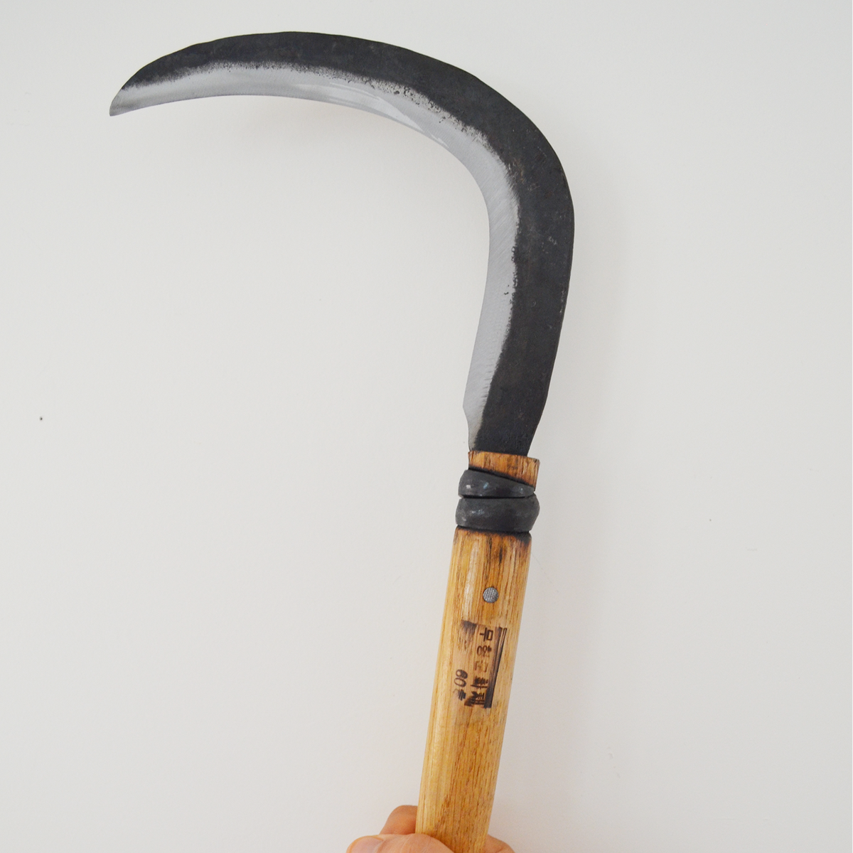 Hand holding a hand-forged large sickle with a curved dark steel blade and light wooden handle, shown against a clean neutral background to highlight its size and artisan design.