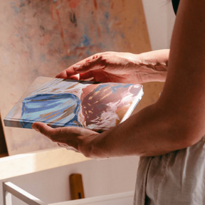 Person holding a small abstract painting in front of a larger artwork.