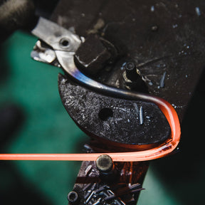A close-up of a blacksmith's anvil with a glowing orange-red heated metal piece being shaped by a hammer, set against a dark, blurred background.