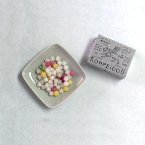 A small square dish filled with multicolored popcorn-like snack pieces next to a silver box labeled KONPEIDOU, photographed on a light neutral background.