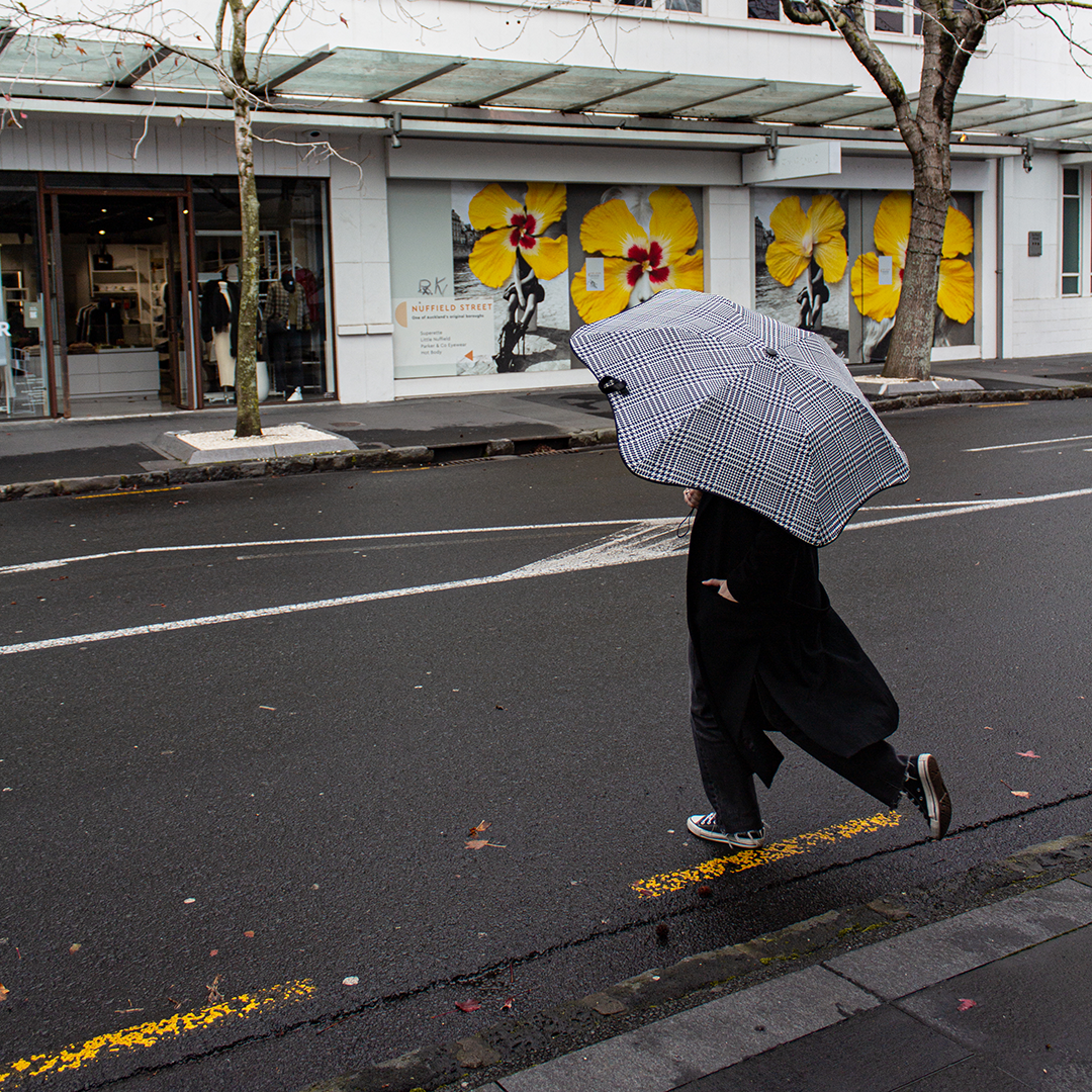 Person holding a checkered umbrella crossing a street with a building featuring large yellow floral murals in the background.