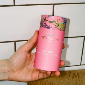Hand holding a pink 'Wild Chai' container with a colorful lid against a tiled wall background.