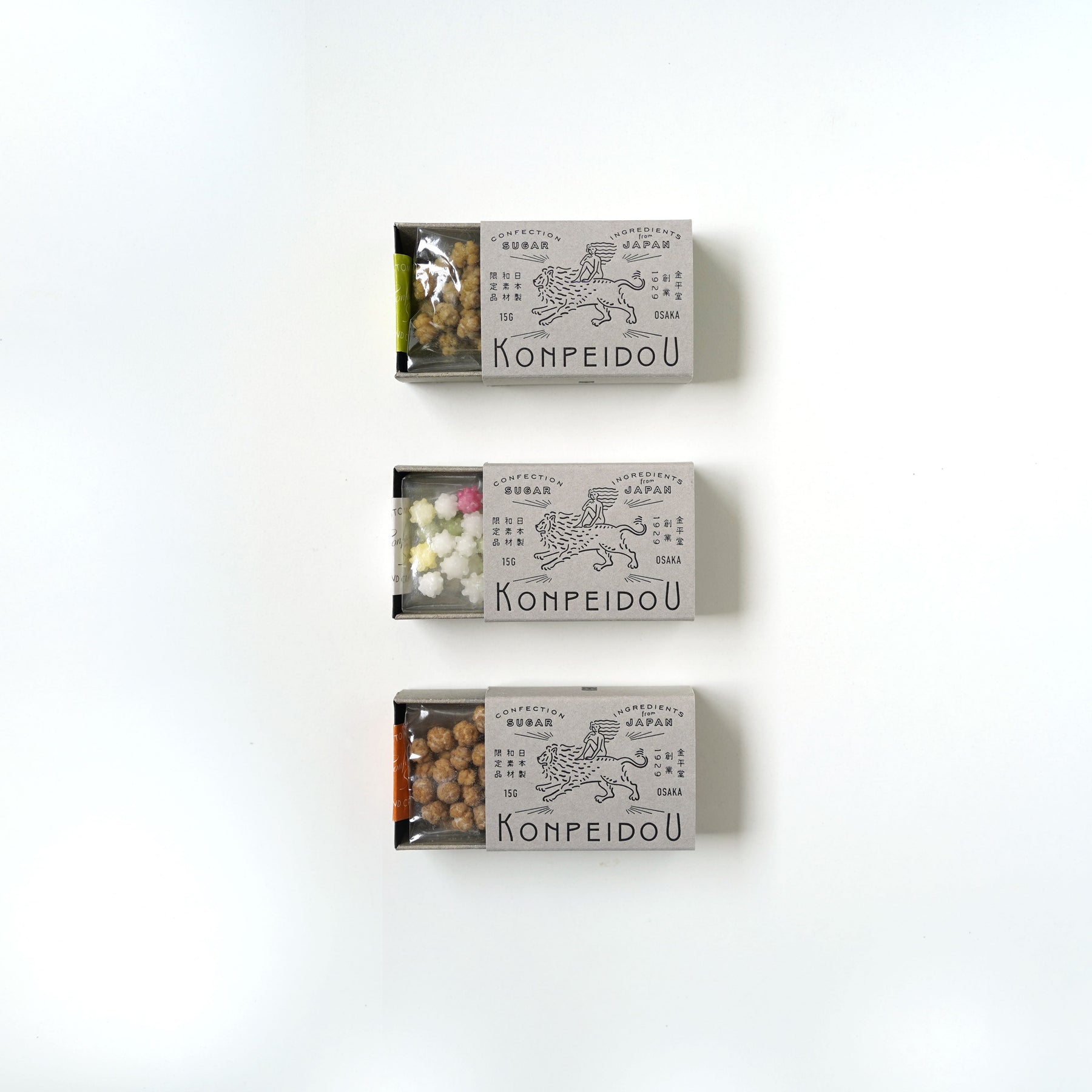 Three small rectangular boxes of Kompeidoó» sweets arranged vertically on a plain white background. The boxes are light gray with black line drawings and text, and the tops are slightly open to reveal the colorful contents inside.