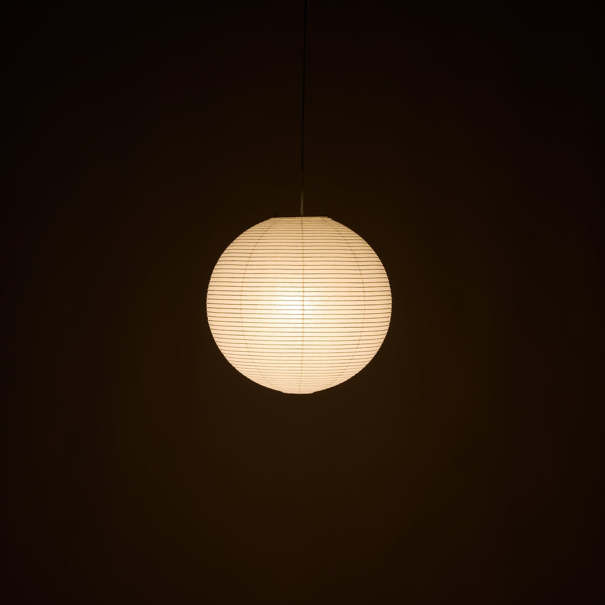 A spherical paper lantern pendant light glowing with warm cream light against a dark background.