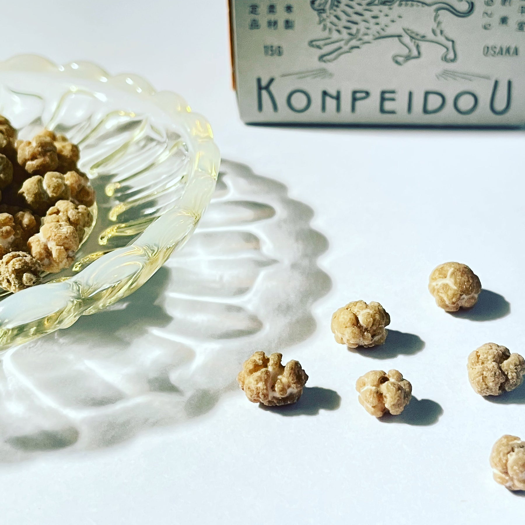 Konpeidou brand dried snail capsules shown with their white and green packaging box and a clear glass dish filled with the brown, textured capsules, set against a bright white background with artistic shadow casting.