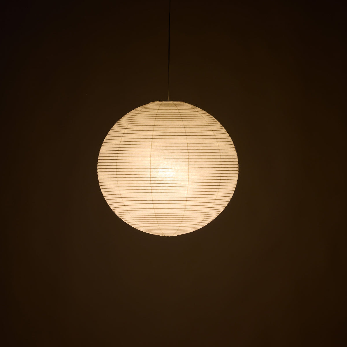 A spherical paper lantern pendant light glowing with warm cream light, suspended by a thin black cord against a dark brown background.