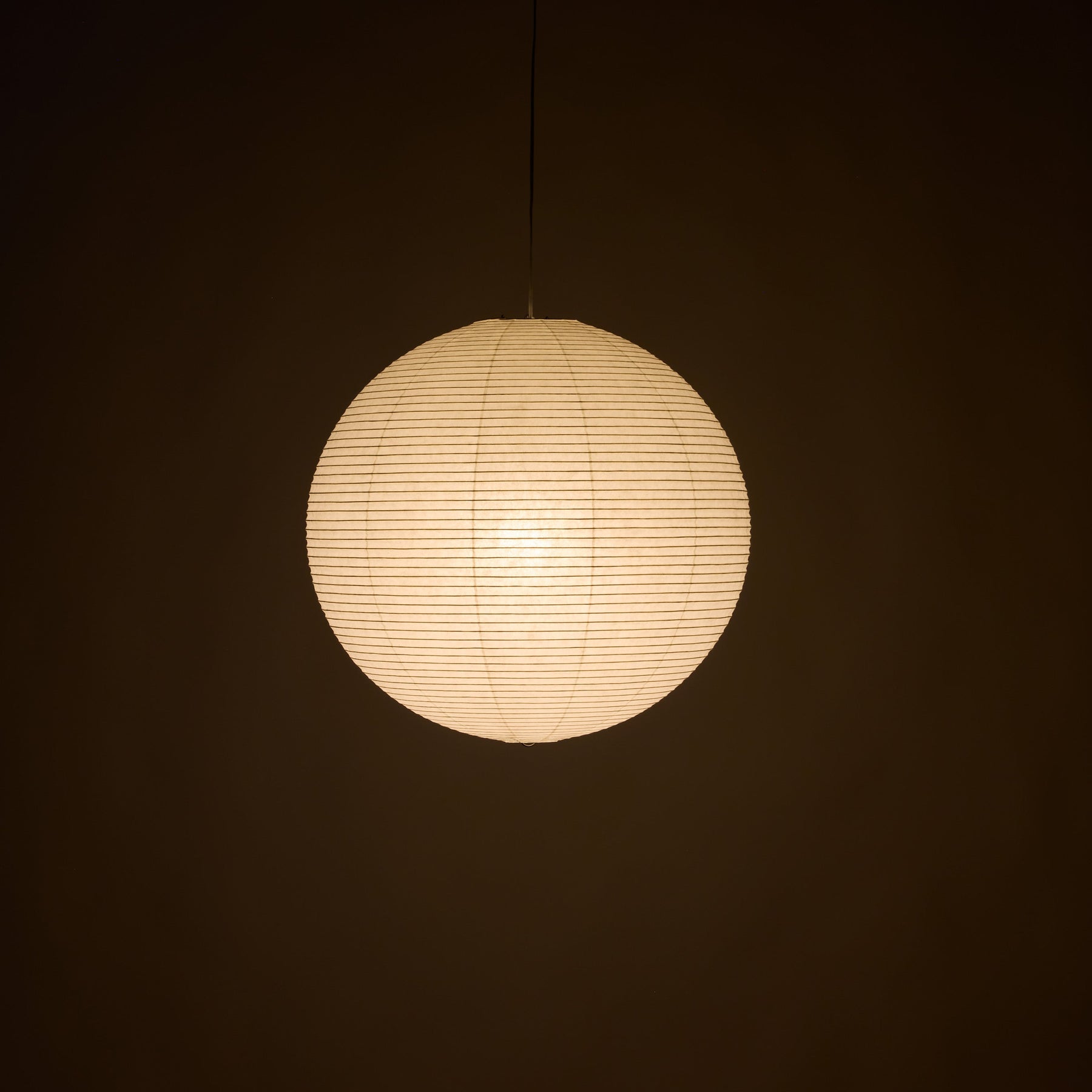 A spherical paper lantern pendant light glowing with warm cream light, suspended by a thin black cord against a dark brown background.