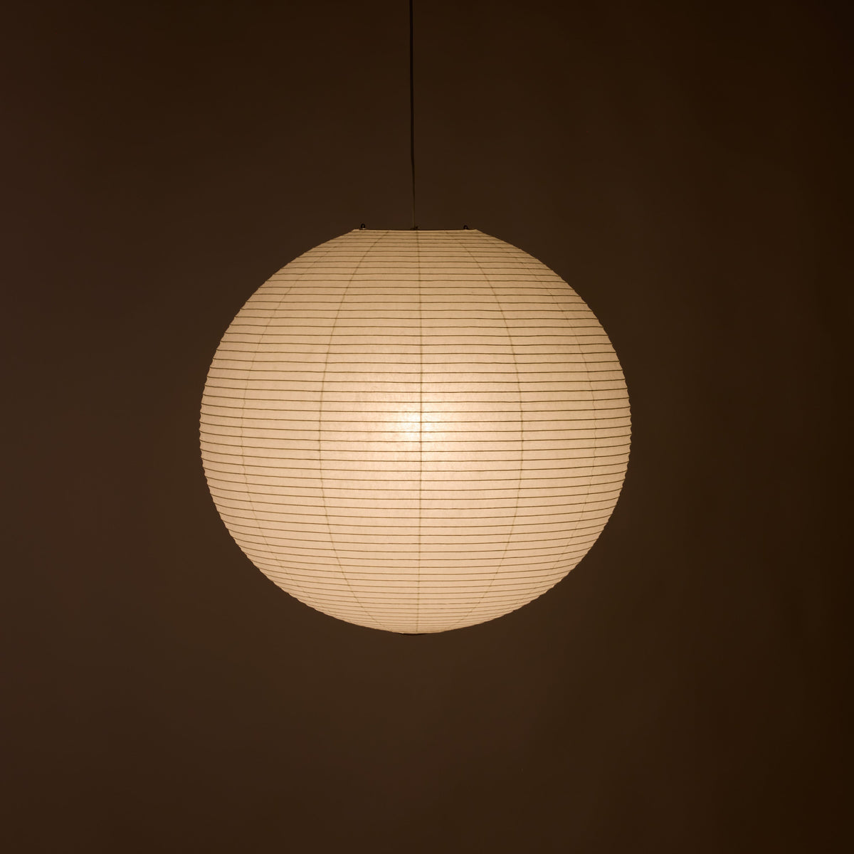 A spherical paper lantern pendant light glowing with warm cream light against a dark brown background.