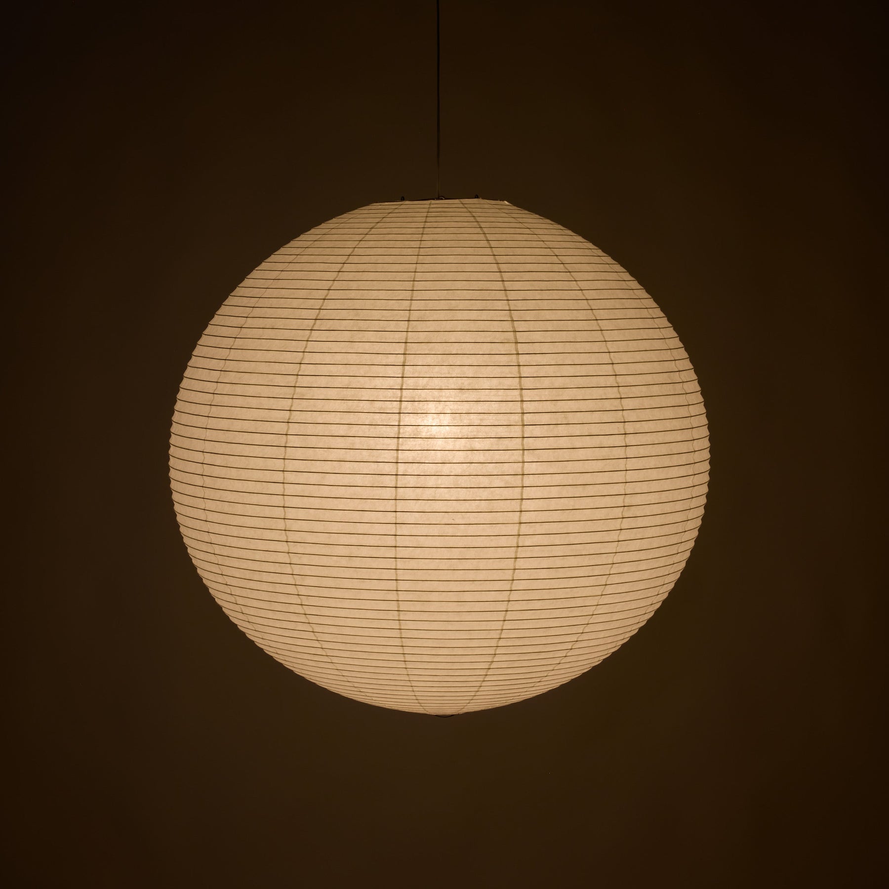 A spherical paper lantern pendant light glowing with warm cream light against a dark brown background.