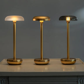 Three modern table lamps with slim gold-finished stems and round bases are displayed on a light-colored console table against a dark gray wall. The left lamp has a frosted white dome shade, the middle lamp has a brushed gold dome shade, and the right lamp has a smoked glass dome shade, all illuminated.