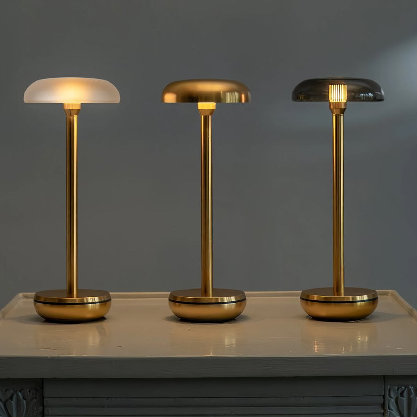 Three modern table lamps with slim gold-finished stems and round bases are displayed on a light-colored console table against a dark gray wall. The left lamp has a frosted white dome shade, the middle lamp has a brushed gold dome shade, and the right lamp has a smoked glass dome shade, all illuminated.