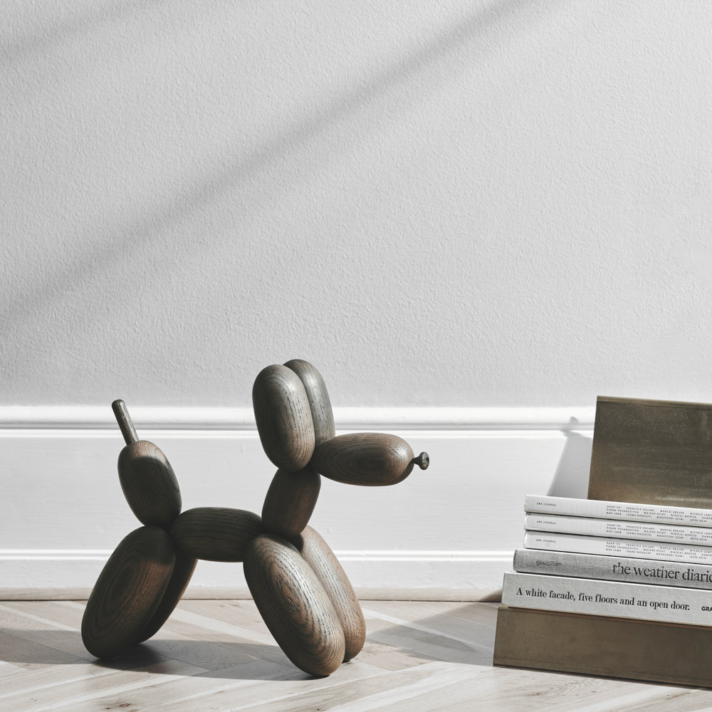 Wooden balloon dog sculpture on a light wooden floor with a stack of books in the background.