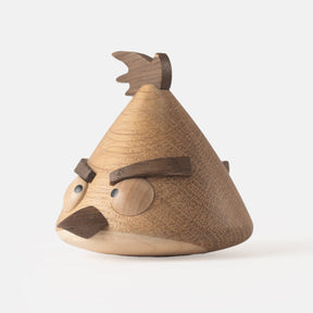 Side view of wooden figurine of an animated cartoon bird character carved from light and dark wood, featuring a triangular body shape, prominent angled eyebrows, round eye details, a small pointed beak, visible natural wood grain, and a smooth matte surface on a plain white background.