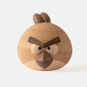 Wooden figurine of an animated cartoon bird character carved from light and dark wood, featuring a round body, prominent angular eyebrows, circular eye details, a pointed beak, visible natural wood grain, and a smooth matte surface on a plain white background.