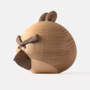 Side view of wooden figurine of an animated cartoon bird character carved from light and dark wood, featuring a round body, prominent angular eyebrows, circular eye details, a pointed beak, visible natural wood grain, and a smooth matte surface on a plain white background.