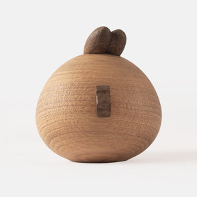 Back view of wooden figurine of an animated cartoon bird character carved from light and dark wood, featuring a round body, prominent angular eyebrows, circular eye details, a pointed beak, visible natural wood grain, and a smooth matte surface on a plain white background.