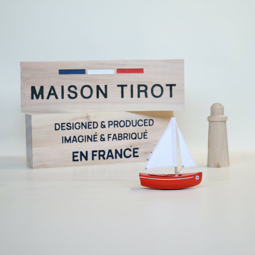 Red sailboat model with a wooden sign displaying 'MAISON TIROT' on a light background