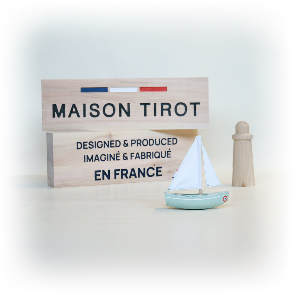 Wooden toy sailboat and lighthouse in front of a 'MAISON TIROT' sign on a light background