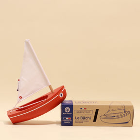 Red toy sailboat with white sail next to its packaging on a beige background