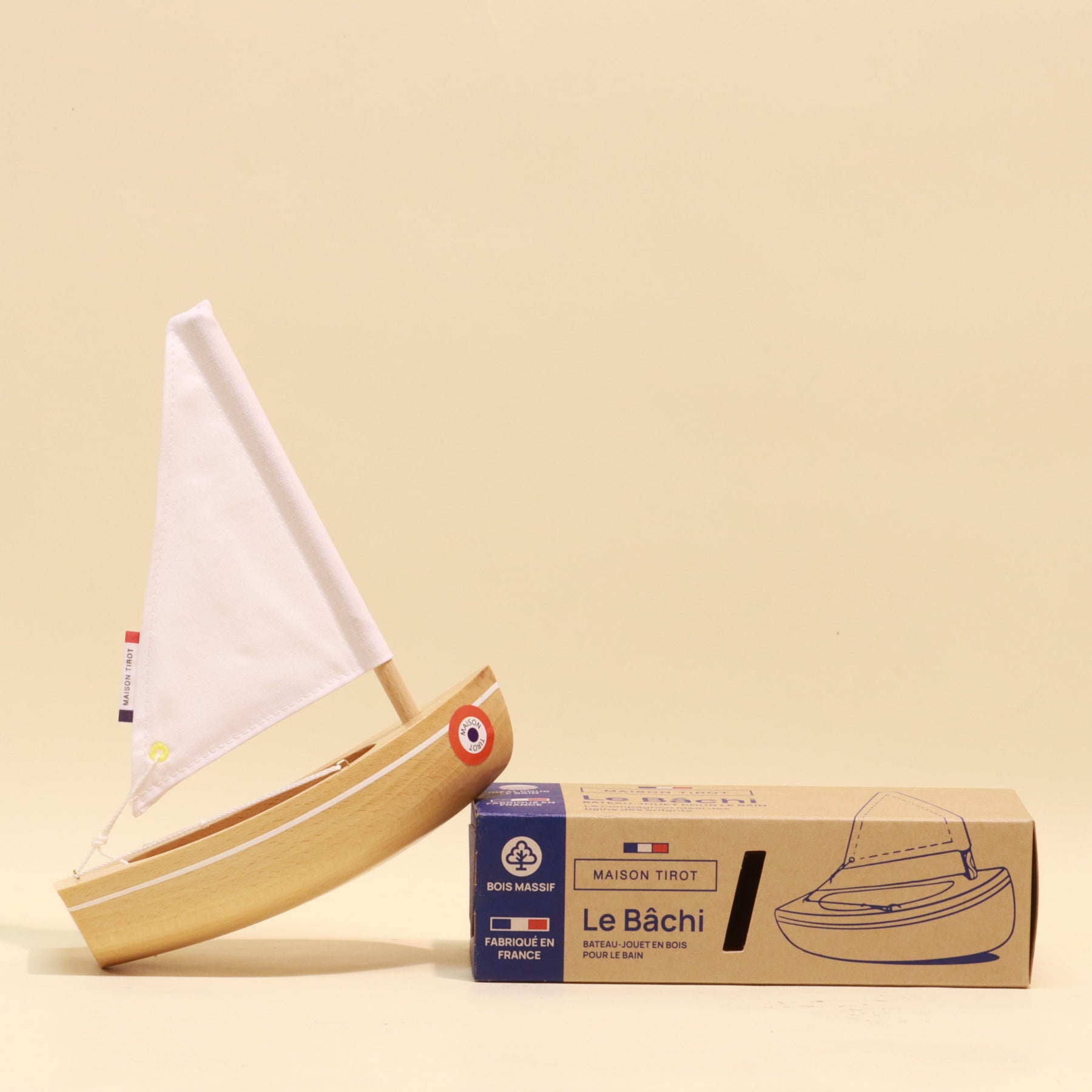 Wooden toy sailboat with a white sail next to its packaging on a beige background