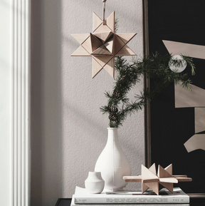 Interior vignette featuring a hanging geometric star ornament crafted from light oak, suspended by a cord above a stack of books and styled with a white ceramic vase and evergreen branch, creating a minimalist, softly lit decorative scene.