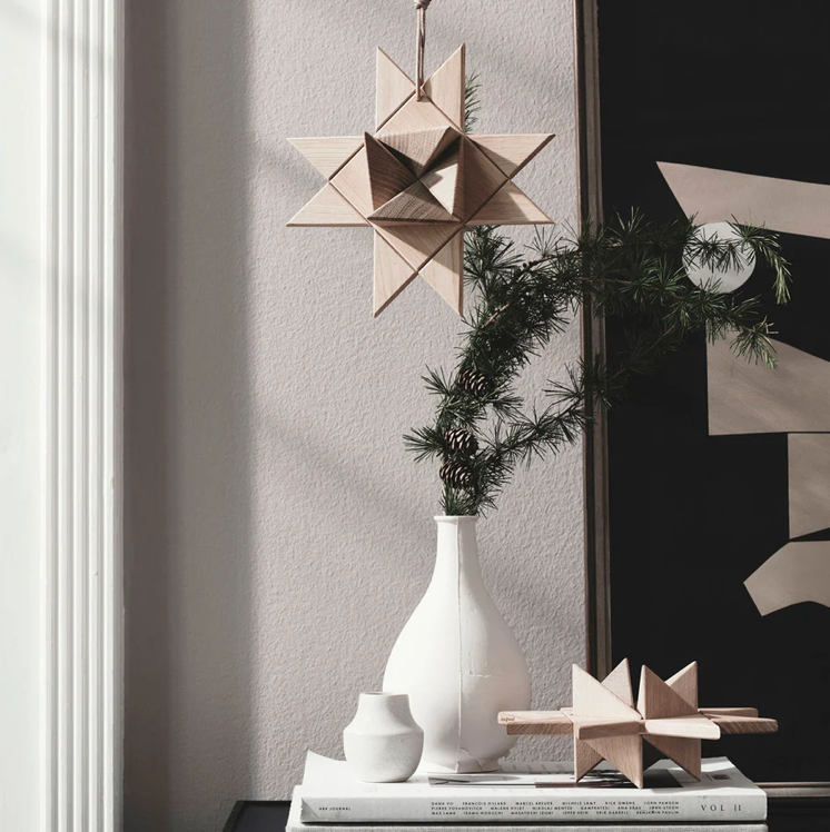 Interior vignette featuring a hanging geometric star ornament crafted from light oak, suspended by a cord above a stack of books and styled with a white ceramic vase and evergreen branch, creating a minimalist, softly lit decorative scene.