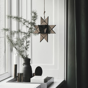 Interior scene featuring a hanging geometric star ornament made from smoked wood, suspended by a cord near a window and styled with minimalist vases, candles, and greenery on a windowsill, creating a calm, modern atmosphere with soft natural light.