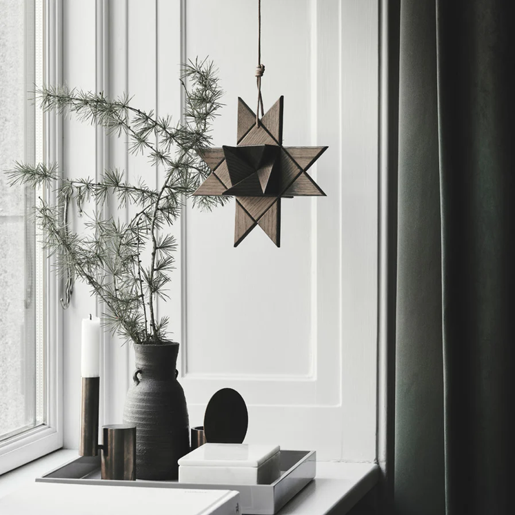 Interior scene featuring a hanging geometric star ornament made from smoked wood, suspended by a cord near a window and styled with minimalist vases, candles, and greenery on a windowsill, creating a calm, modern atmosphere with soft natural light.