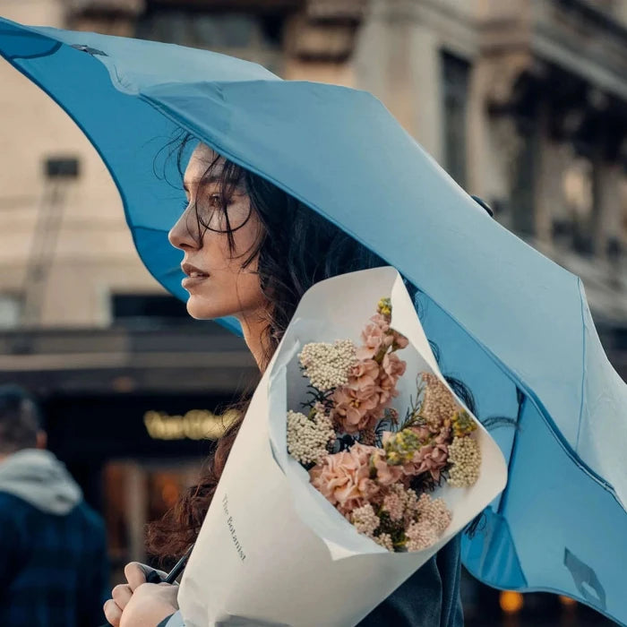 Woman holding a blue umbrella and a bouquet of flowers in an urban setting