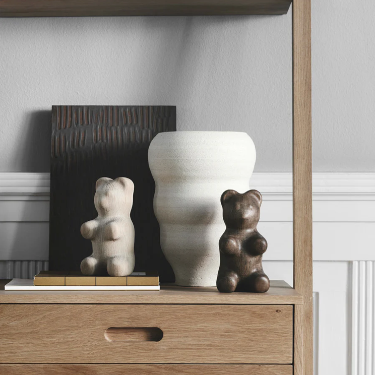 Interior shelf styling featuring two small bear-shaped wooden figurines in light and dark tones beside a sculptural white ceramic vase, arranged on stacked books within a natural wood shelving unit against a soft gray wall.