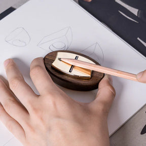 Hand using a wooden pencil sharpener with a pencil, on a white surface with sketches.