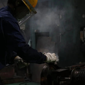 A worker in a dark blue uniform, white gloves, and a yellow hard hat handles a glowing orange-red piece of molten metal in an industrial setting with smoke and machinery.