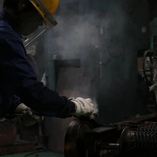 A worker in a dark blue uniform, white gloves, and a yellow hard hat handles a glowing orange-red piece of molten metal in an industrial setting with smoke and machinery.