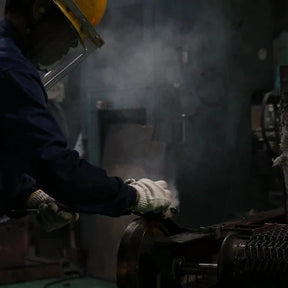 A worker in a dark blue uniform, white gloves, and a yellow hard hat handles a glowing orange-red piece of molten metal in an industrial setting with smoke and machinery.
