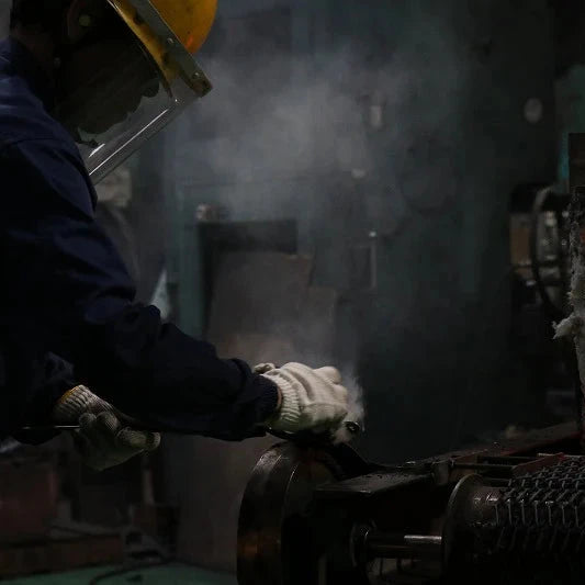 A worker in a dark blue uniform, white gloves, and a yellow hard hat handles a glowing orange-red piece of molten metal in an industrial setting with smoke and machinery.