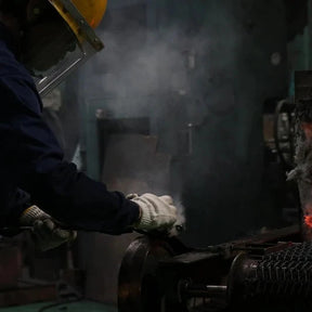 A worker in a dark blue uniform, white gloves, and a yellow hard hat handles a glowing orange-red piece of molten metal in an industrial setting with smoke and machinery.