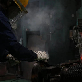 A worker in a dark blue uniform, white gloves, and a yellow hard hat handles a glowing orange-red piece of molten metal in an industrial setting with smoke and machinery.