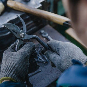Person wearing gloves holding a pair of shears over a workbench with tools in the background