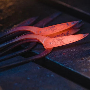 A close-up of glowing hot metal tools or bars resting on a dark workbench, with one piece illuminated in bright orange-red heat.