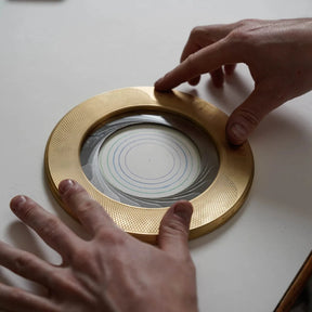 Gold circular object with a reflective surface, held by two hands on a light background