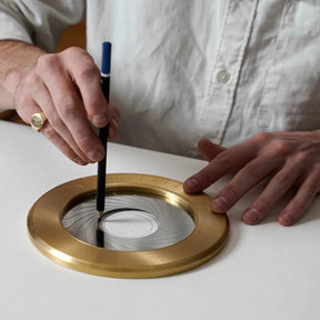 Person using a pen on a gold circular object with concentric circles on a white surface
