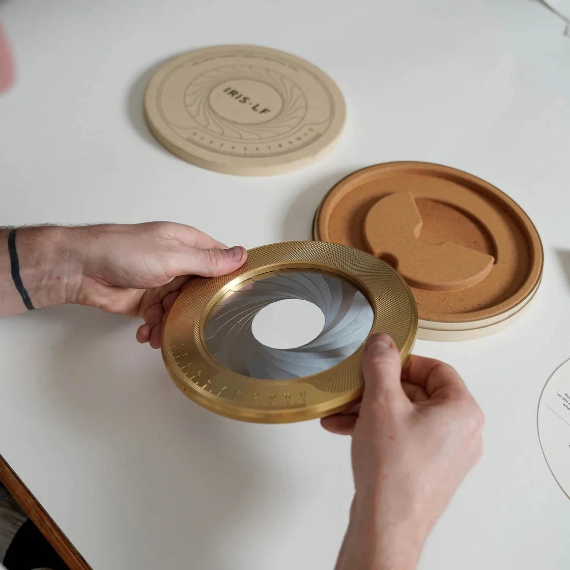 Hands holding a precision-machined brass iris aperture tool with engraved measurement markings, shown open to reveal overlapping metal blades, with its cork-lined case and labeled lid on a white work surface.