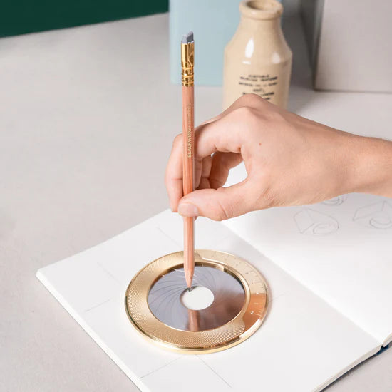 Hand using a gold pen to draw on a white surface with a circular design.