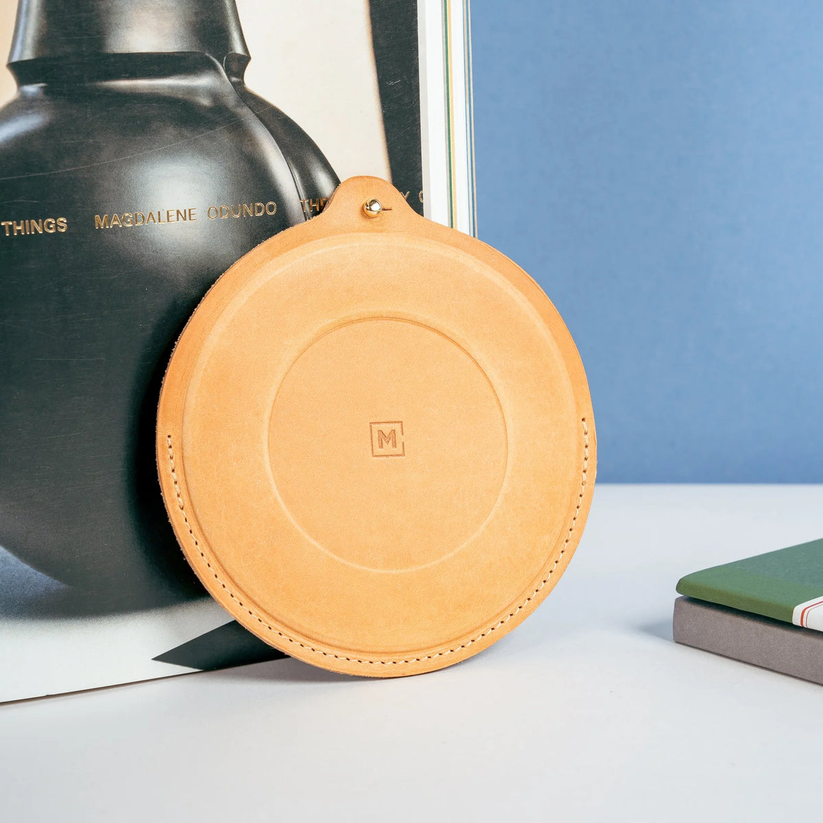 Round tan leather sheath with hand-stitched edges and a small brass rivet, displayed upright against an art book and blue backdrop on a clean tabletop.