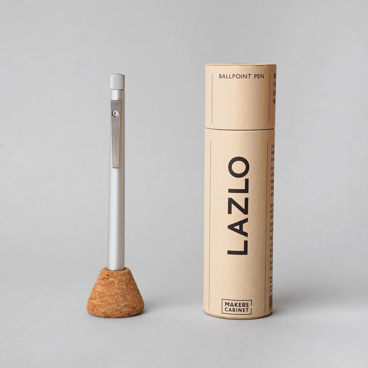 Minimalist aluminum ballpoint pen standing upright in a cork base beside a cylindrical cardboard tube labeled ‘LAZLO,’ showcasing a clean, modern writing tool and its sustainable packaging.