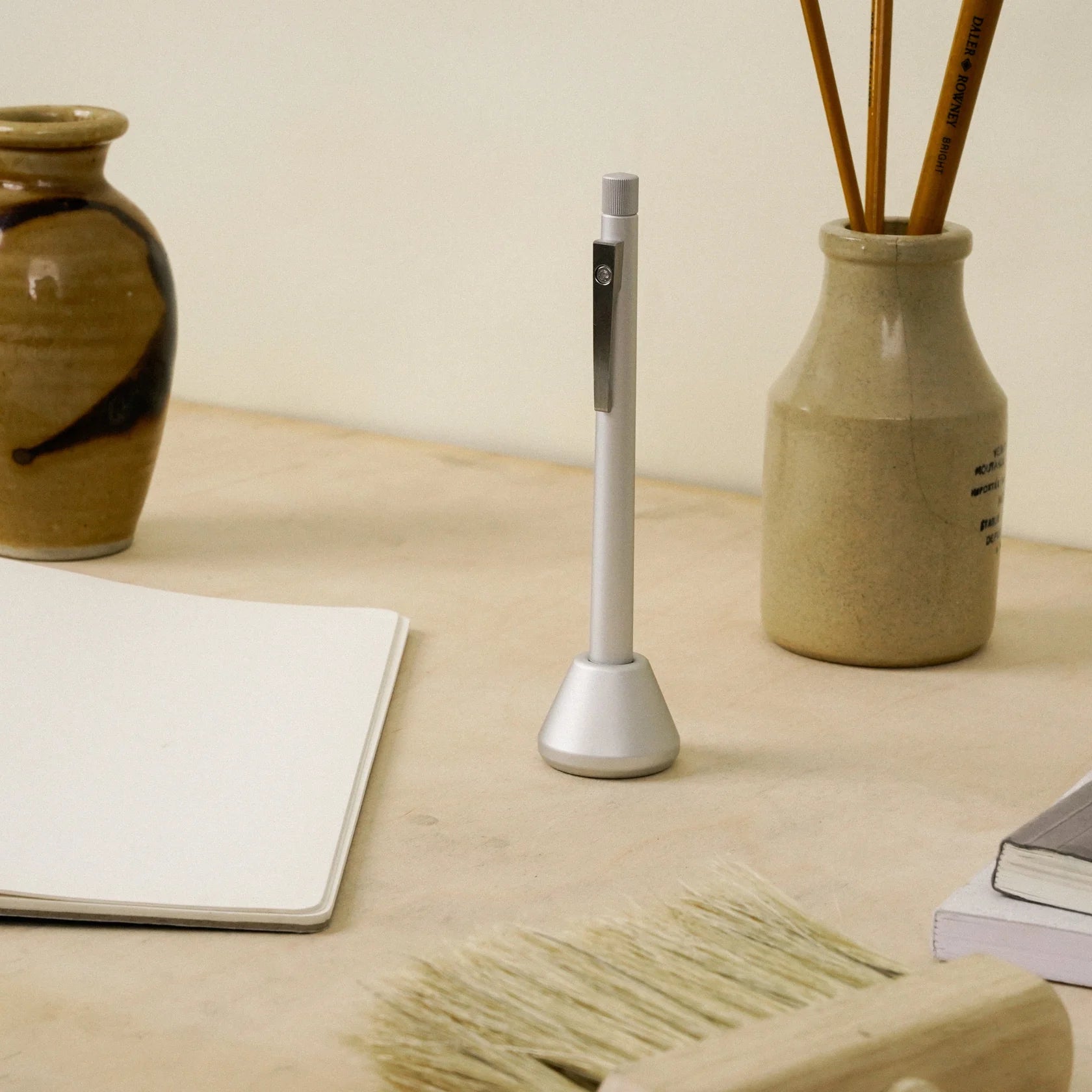 Minimalist aluminum ballpoint pen standing upright in a matching aluminum desk stand on a light wood workspace, surrounded by a notebook, ceramic jars with pencils, and simple studio accessories.