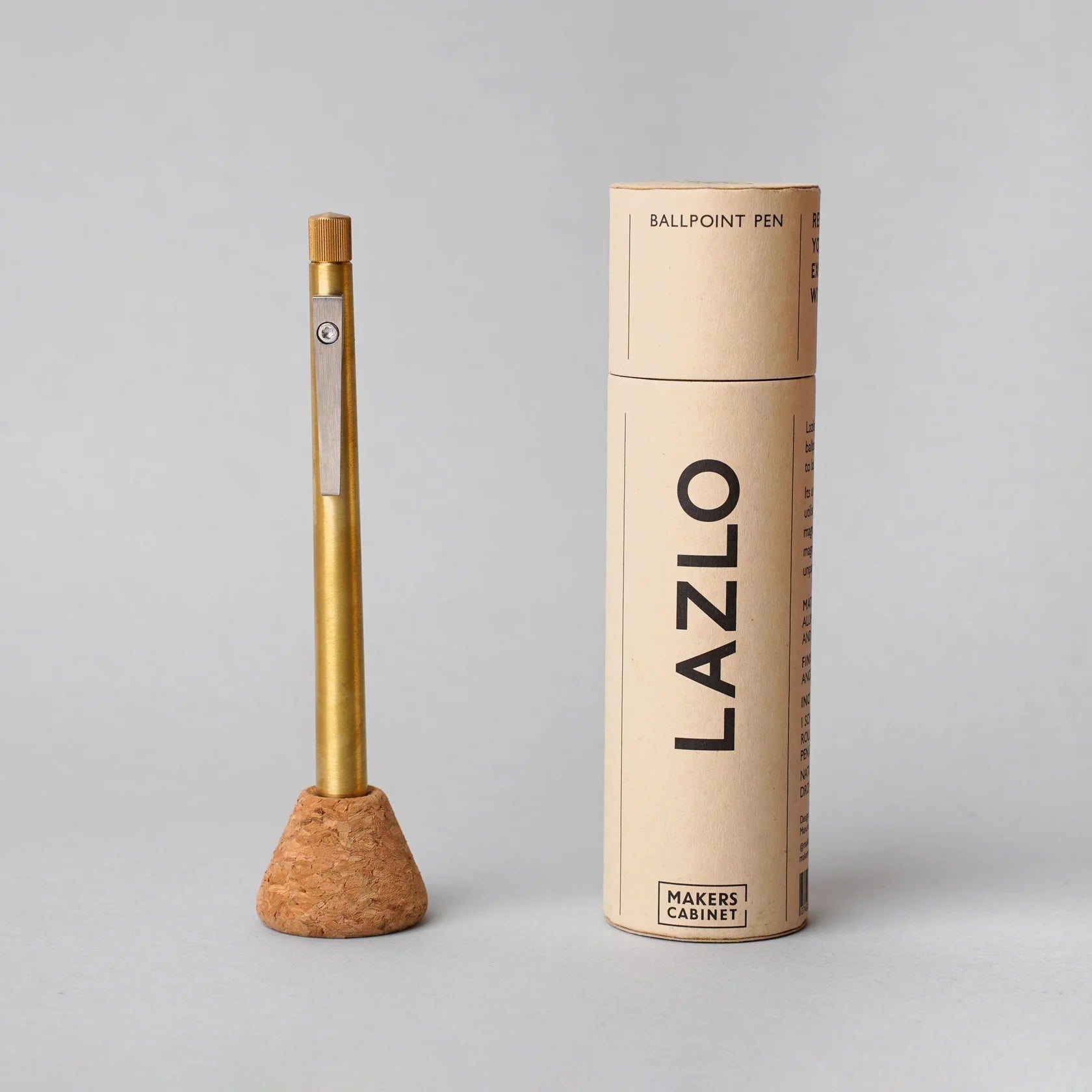 Brass ballpoint pen displayed upright in a cork desk stand beside a cylindrical cardboard packaging tube labeled ‘LAZLO,’ highlighting a minimalist writing instrument with a brushed metal finish against a neutral gray background.