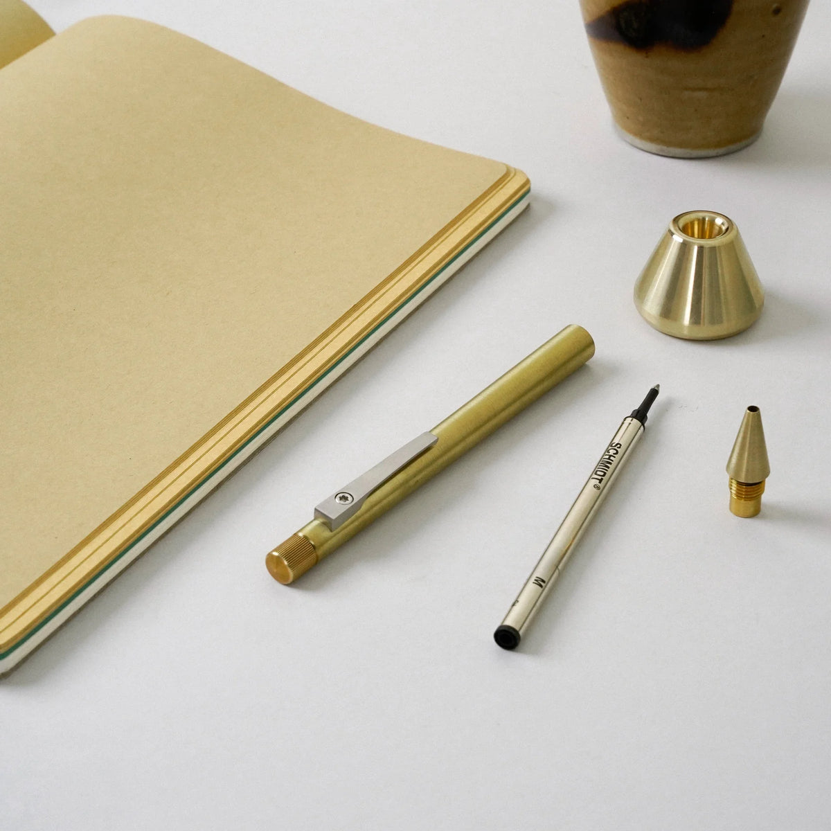 Minimal desk flat lay featuring a brass pen with a removable ink refill and interchangeable brass tips, arranged beside a cream-colored notebook and a sculptural brass pen stand on a clean white surface, highlighting refined materials and functional design.